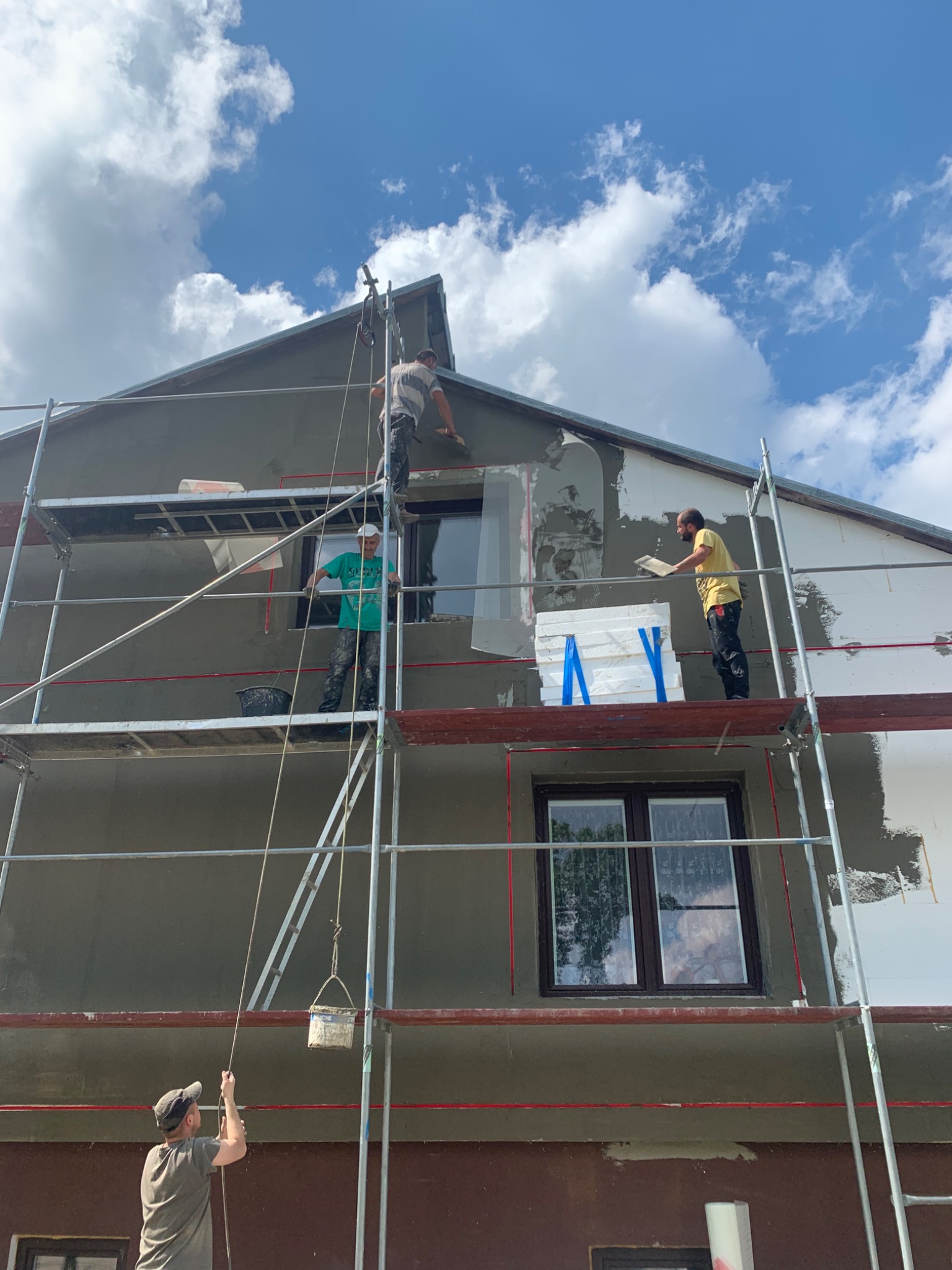 Elewacja domu w trakcie szpachlowania i ocieplania, widoczne rusztowania, pracownicy w trakcie pracy, styropianowe płyty z niebieską taśmą, szpachlowana ściana pod błękitnym niebem.