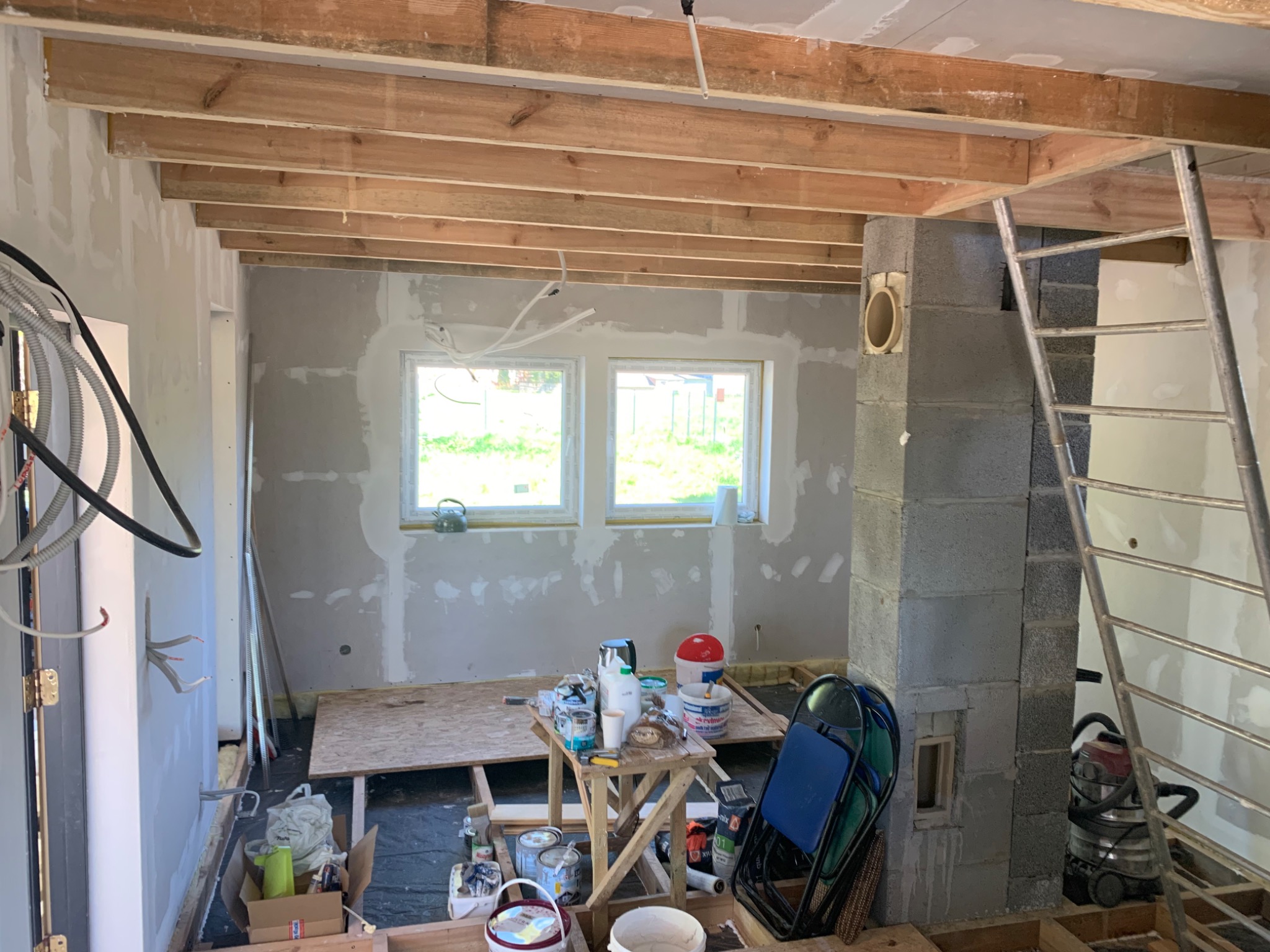 Wnętrze w trakcie prac wykończeniowych: ściany po szpachlowaniu z widocznymi łatami, drewniana konstrukcja sufitu, narzędzia i materiały budowlane na podłodze, drabina oparta o komin z betonu...