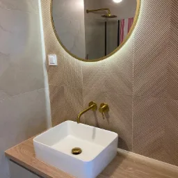 Nowoczesna łazienka z umywalką nablatową, złotą armaturą i okrągłym lustrem z podświetleniem LED. W tle widoczny prysznic z deszczownicą.