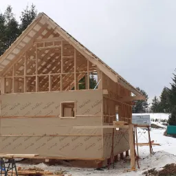 Surowa konstrukcja drewniana domu w trakcie budowy, pokryta membraną izolacyjną Strotex, widoczna na tle zimowego krajobrazu z lasem iglastym.