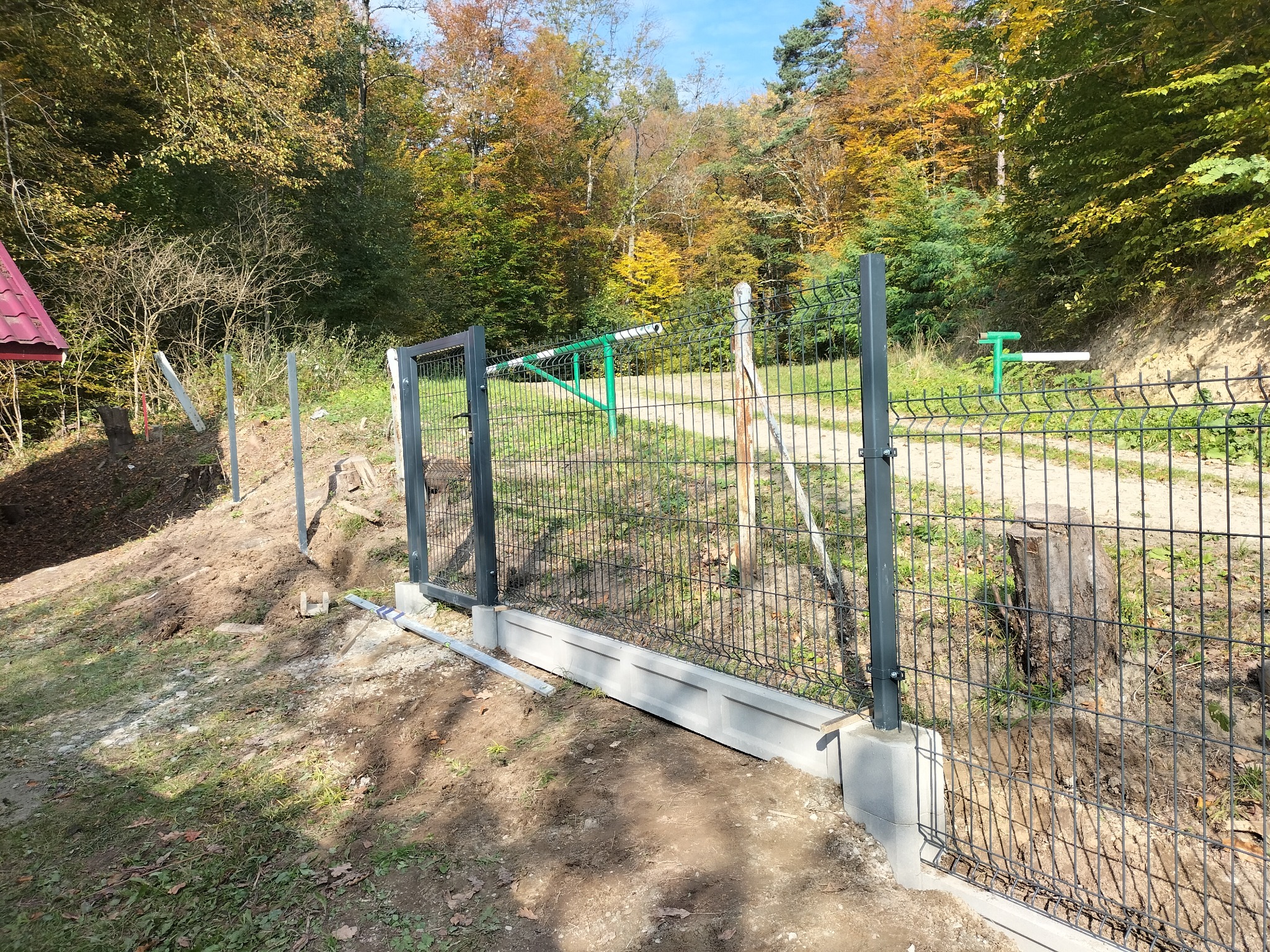 Montaż segmentowej bramy wjazdowej z szarej siatki na betonowej podmurówce, widok na las w jesiennych barwach.