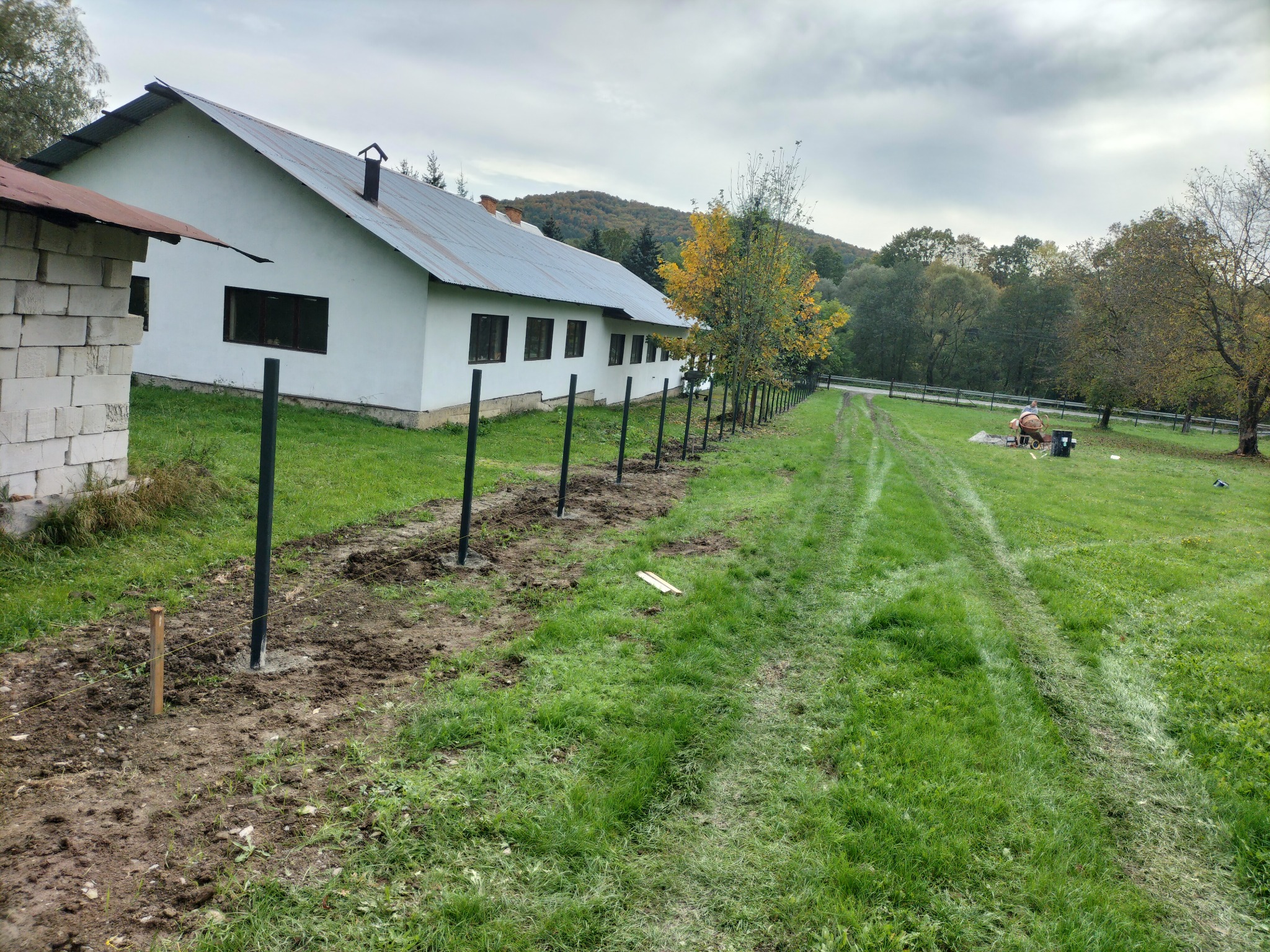Montaż słupków ogrodzeniowych na działce z widokiem na budynek i las, widoczna niwelacja terenu i przygotowanie do betonowania.