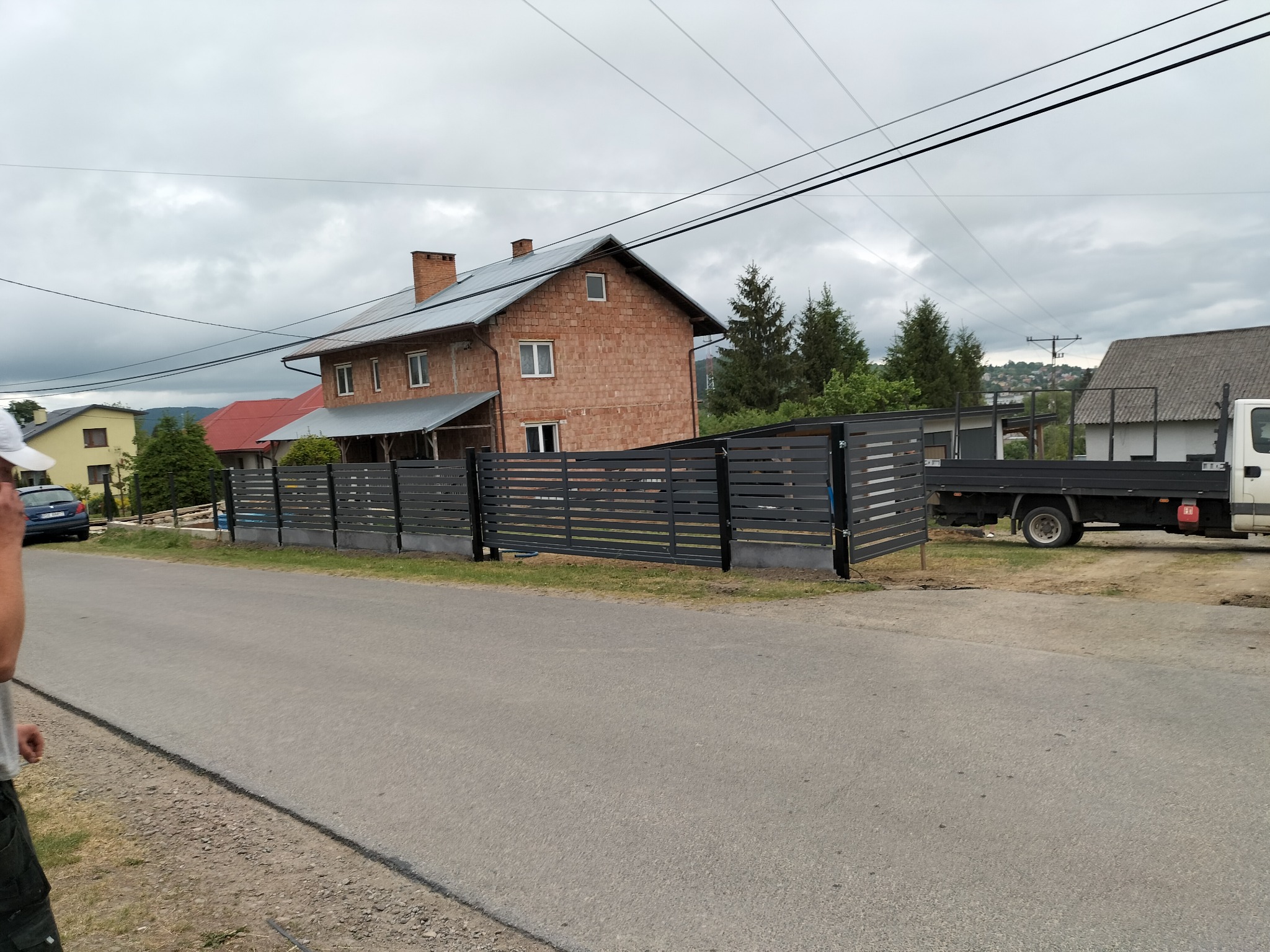 Nowoczesne, poziome przęsła ogrodzeniowe z grafitowej stali, zamontowane przed domem z czerwonej cegły, widoczne z ulicy w pochmurny dzień, z samochodem i ciężarówką w tle.