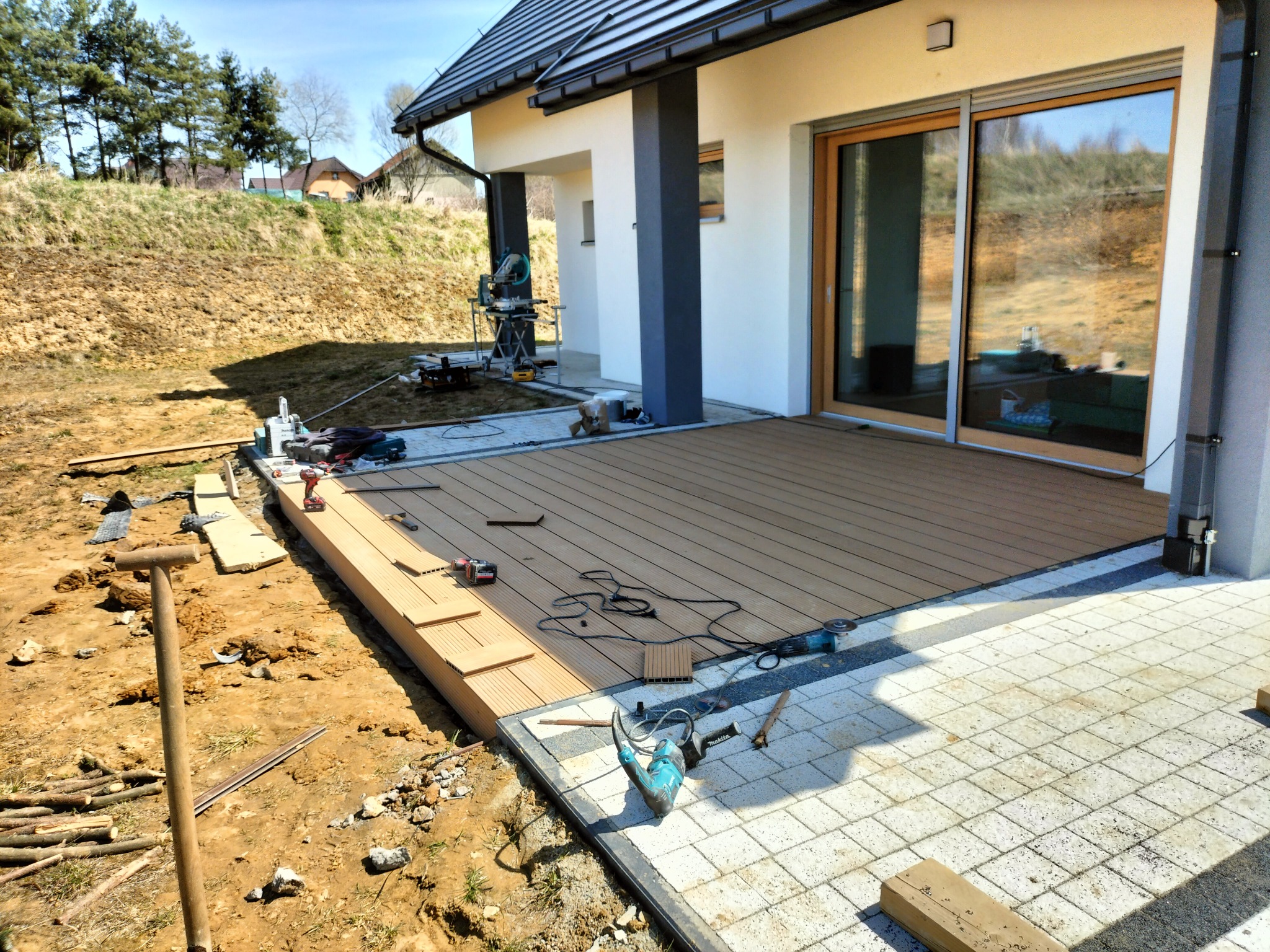 Montaż kompozytowej deski tarasowej na zewnątrz domu jednorodzinnego, widoczne narzędzia, fragment ułożonej nawierzchni z kostki brukowej, ziemia i trawa w tle.