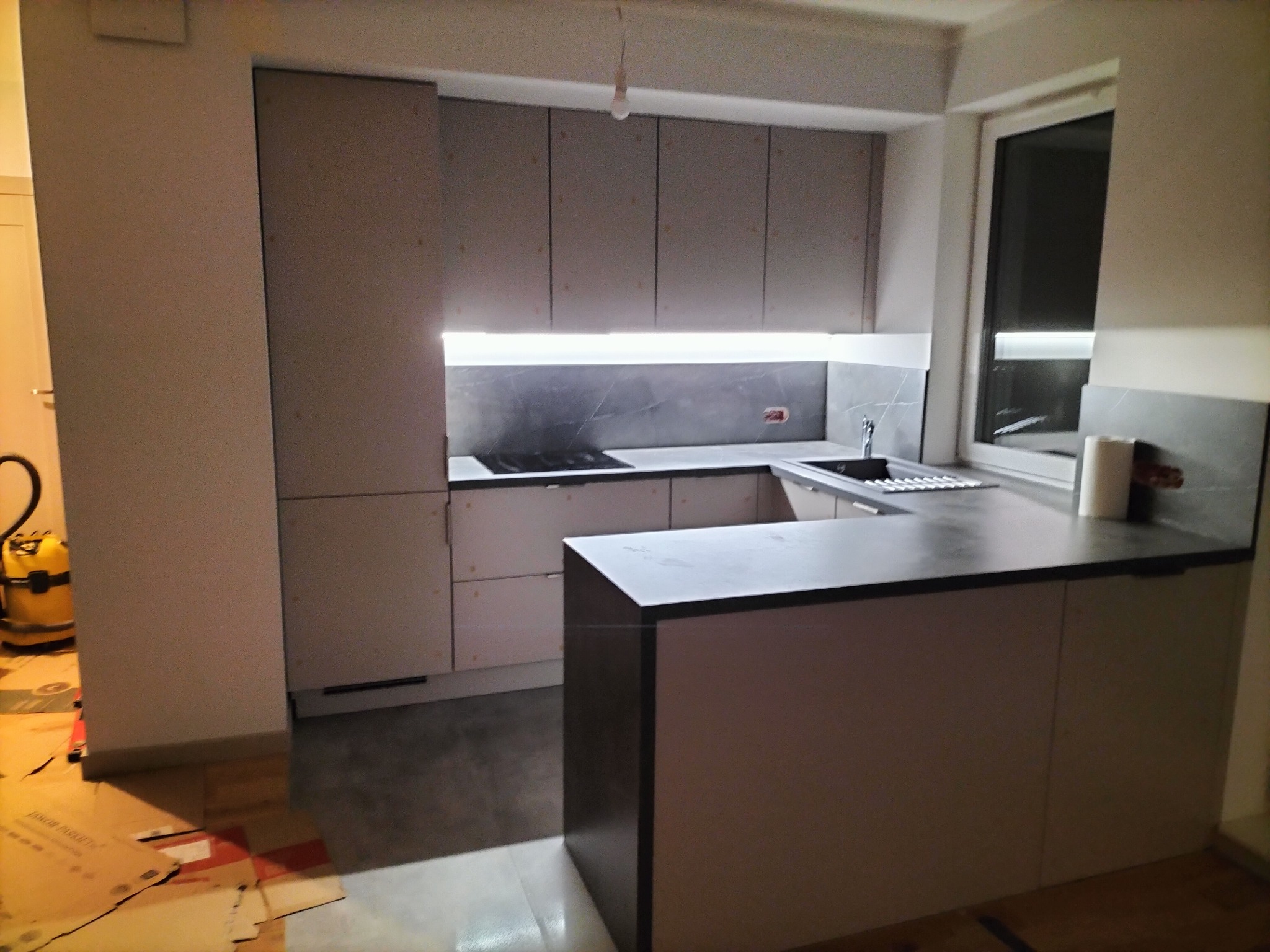 Nowoczesna, jasna kuchnia w trakcie montażu, widoczna zabudowa meblowa z szarymi frontami, czarny blat, oświetlenie LED pod szafkami wiszącymi, kartony na podłodze i odkurzacz przemysłowy w rogu.