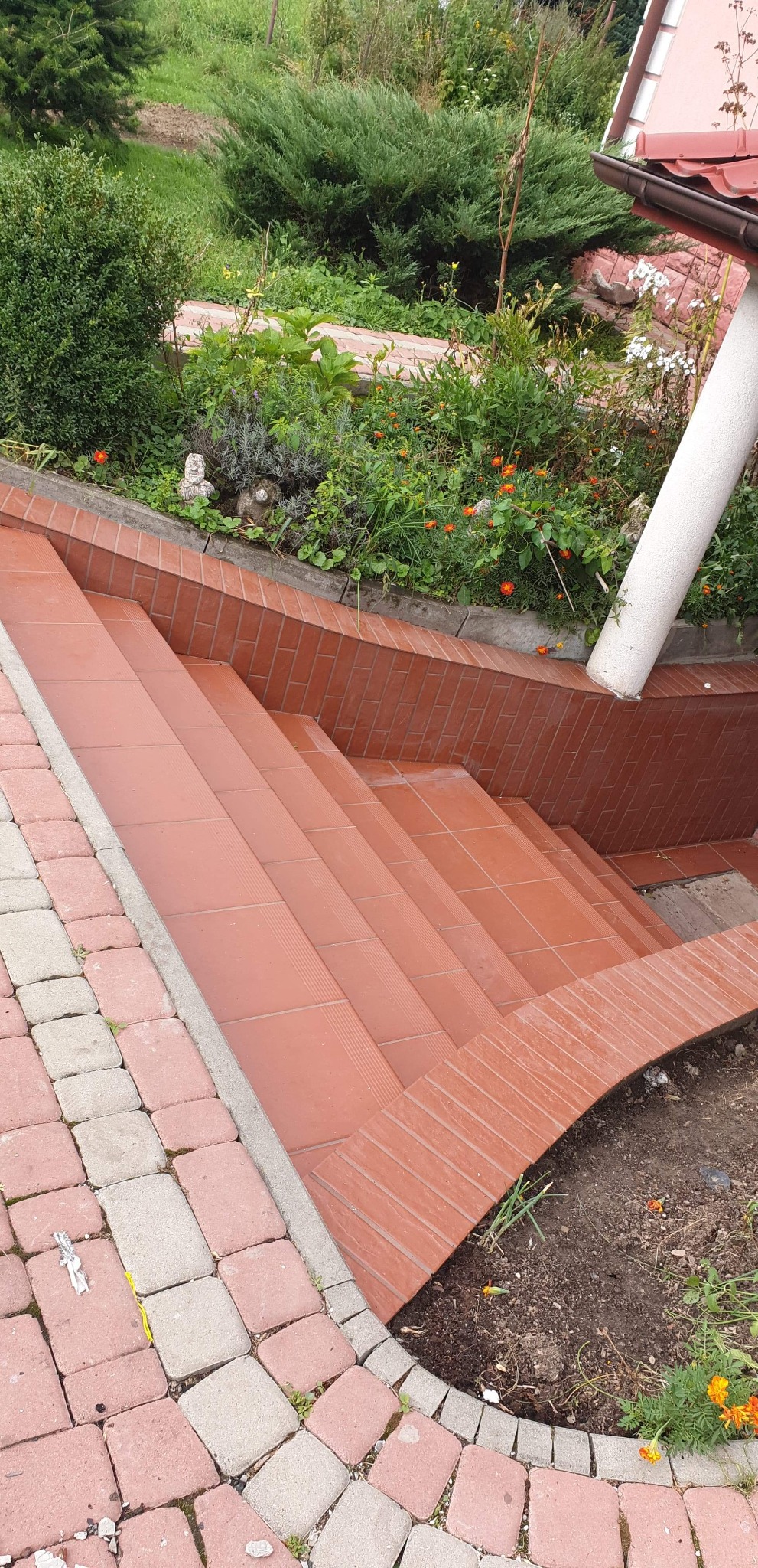 Schody zewnętrzne wyłożone terakotą, z obrzeżem z kostki brukowej, prowadzące do ogrodu z roślinnością i dekoracyjnymi figurkami.