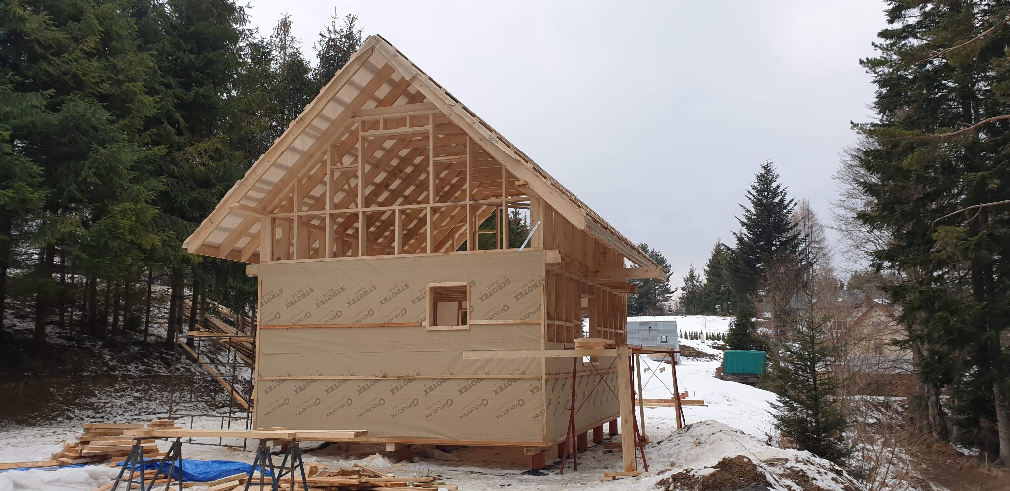 Surowa konstrukcja drewniana domu w trakcie budowy, pokryta membraną izolacyjną Strotex, widoczna na tle zimowego krajobrazu z lasem iglastym.