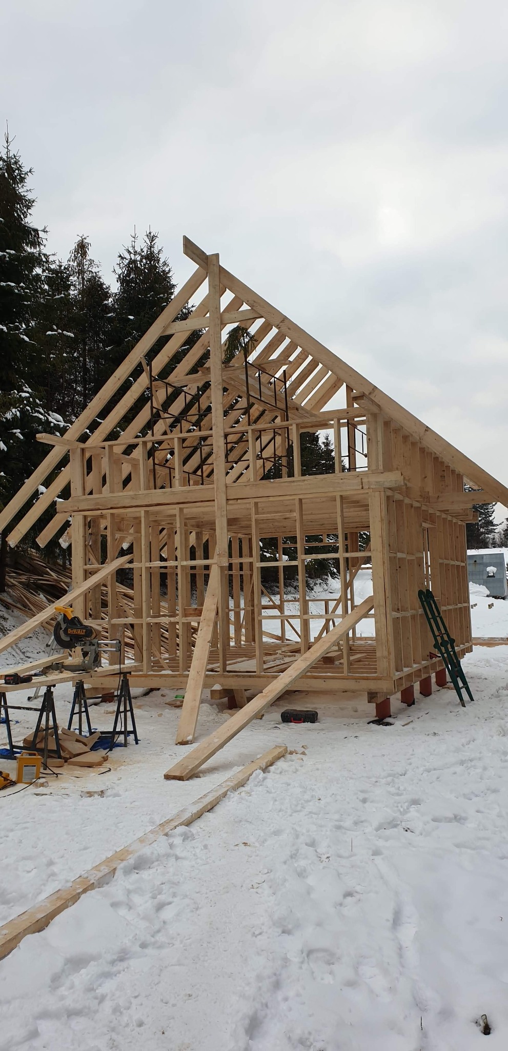 Surowy szkielet drewnianego domu w trakcie budowy, widoczne elementy konstrukcyjne dachu i ścian, piła stołowa DeWalt na statywie, zimowy krajobraz z pokrywą śnieżną i drzewami w tle.