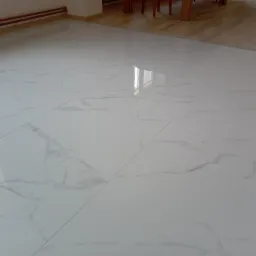 Błyszcząca, biała podłoga z płytek imitujących marmur, z widocznym odbiciem okna i fragmentami drewnianego parkietu oraz grzejnika w tle.