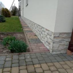 Elewacja budynku z ociepleniem przyziemia imitującym kamień, brązowa rynna spustowa, kolorowa kostka brukowa i rabata z zielonymi roślinami.