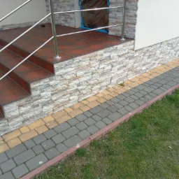 Zewnętrzne schody wejściowe wyłożone brązowymi płytkami, z chromowanymi poręczami, obłożone kamieniem dekoracyjnym, z kostką brukową w odcieniach szarości i żółci oraz trawnikiem.