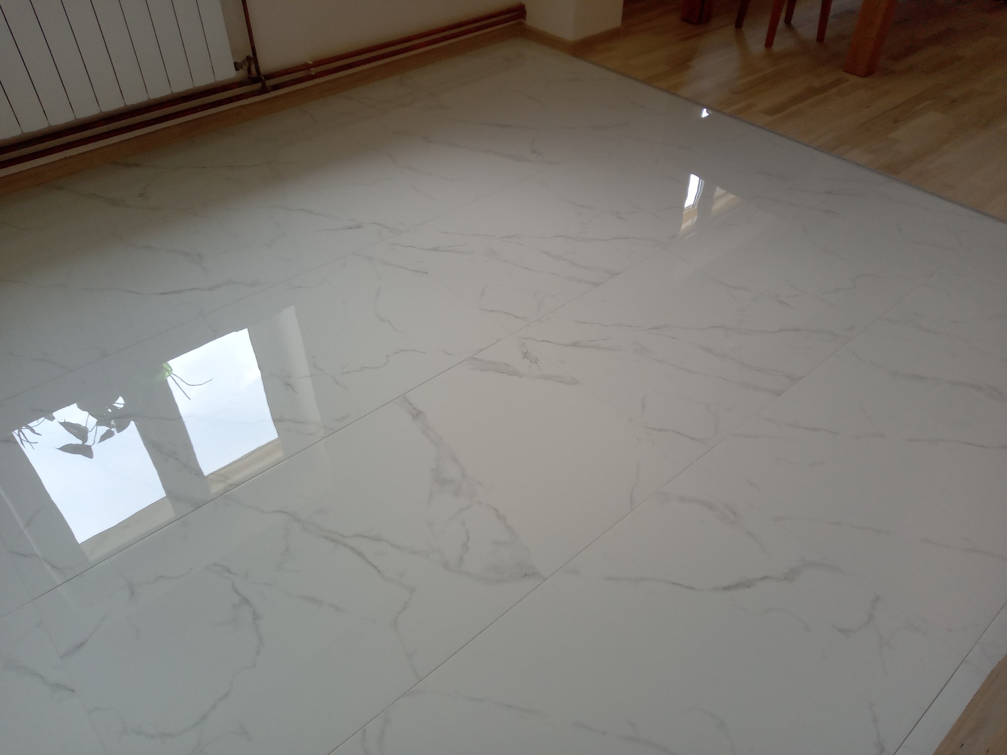 Błyszcząca, biała podłoga z marmurowym wzorem, widoczny grzejnik i fragment drewnianej podłogi w rogu.