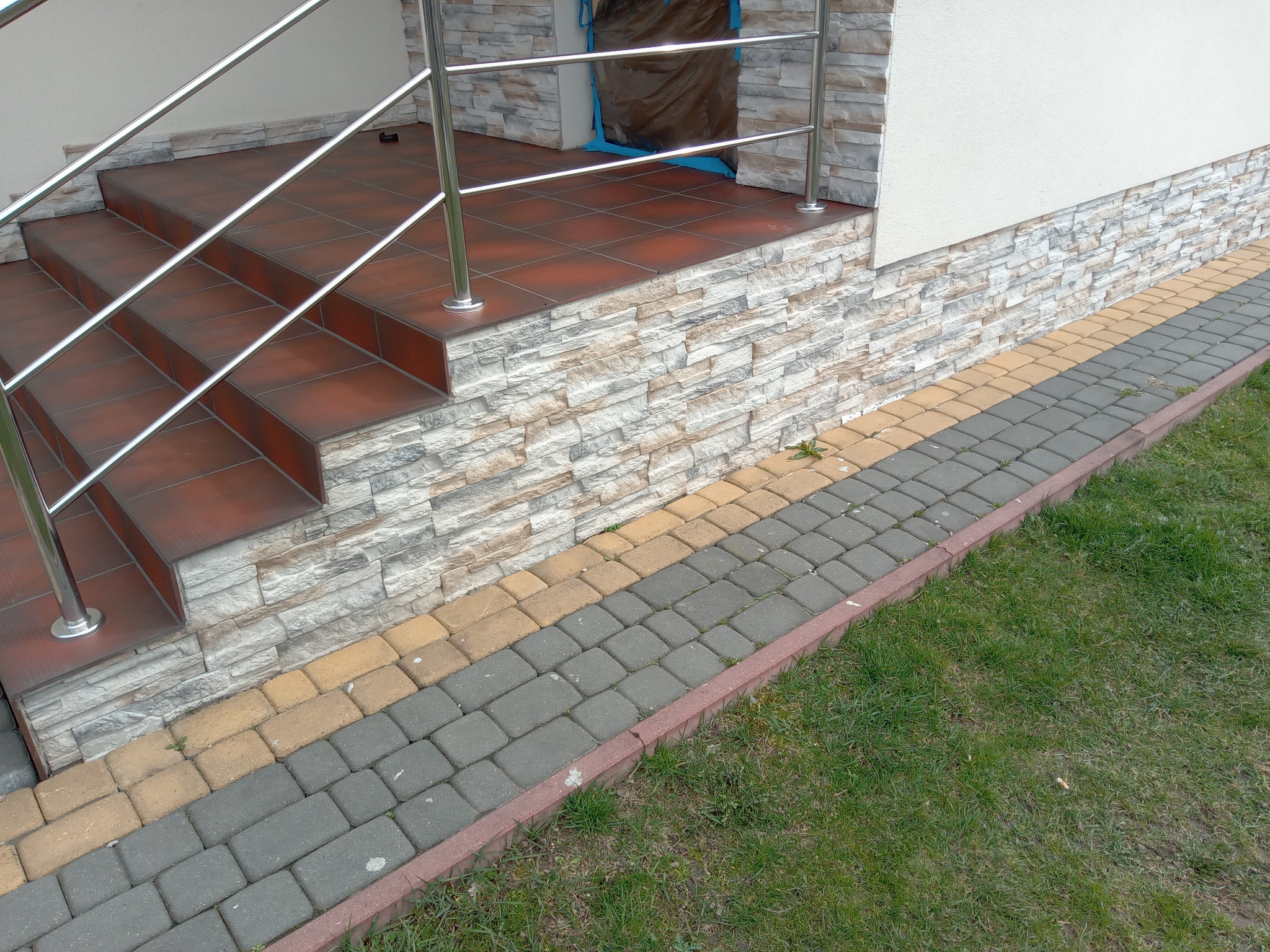 Zewnętrzne schody wejściowe wyłożone brązowymi płytkami, z chromowanymi poręczami, obłożone kamieniem dekoracyjnym, z kostką brukową w odcieniach szarości i żółci oraz trawnikiem.