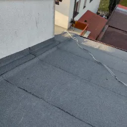 Płaski dach pokryty papą z widocznymi łączeniami, fragment ściany budynku i przewody elektryczne na dachu.