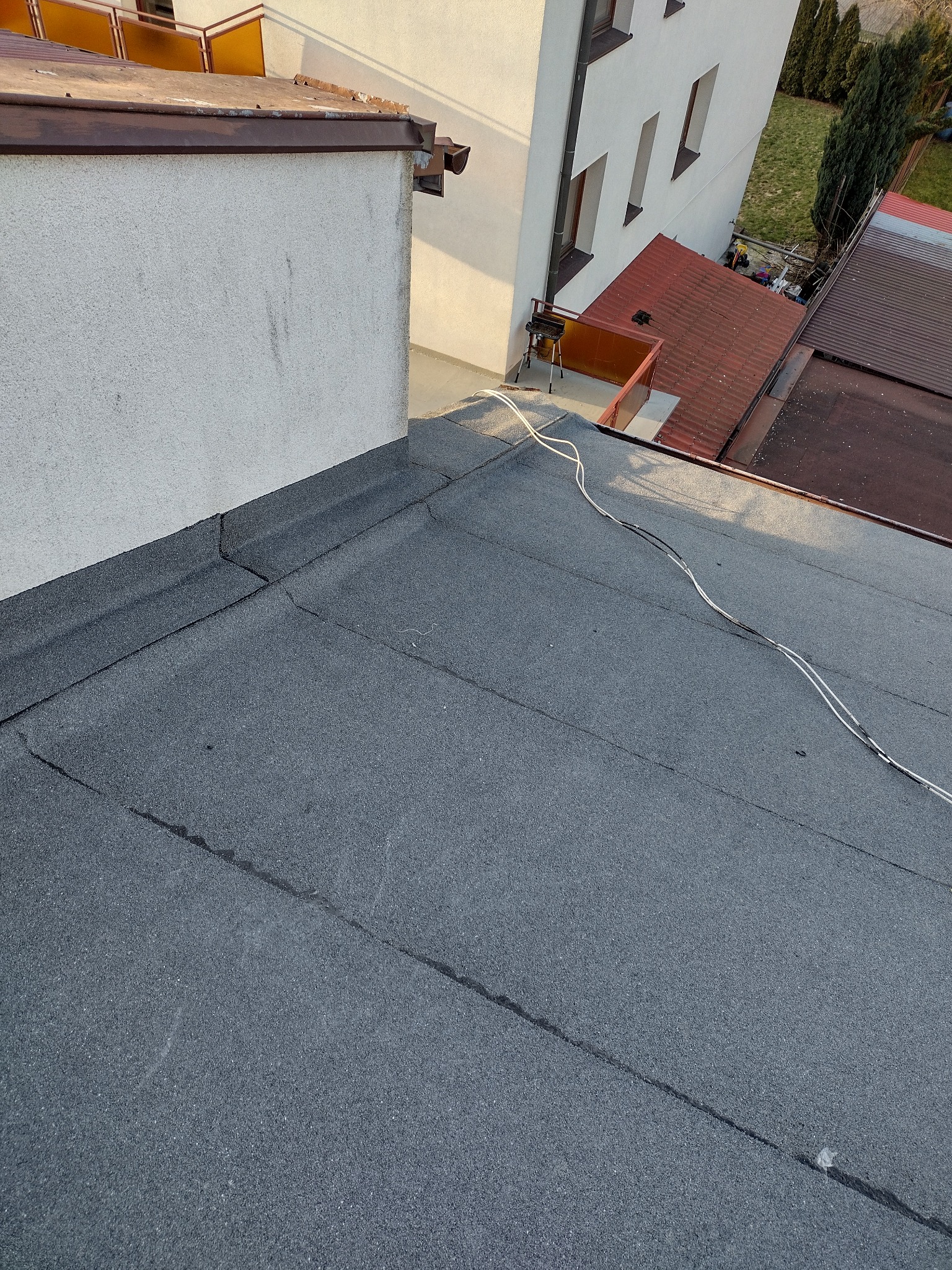Płaski dach pokryty papą z widocznymi łączeniami, fragment ściany budynku i przewody elektryczne na dachu.