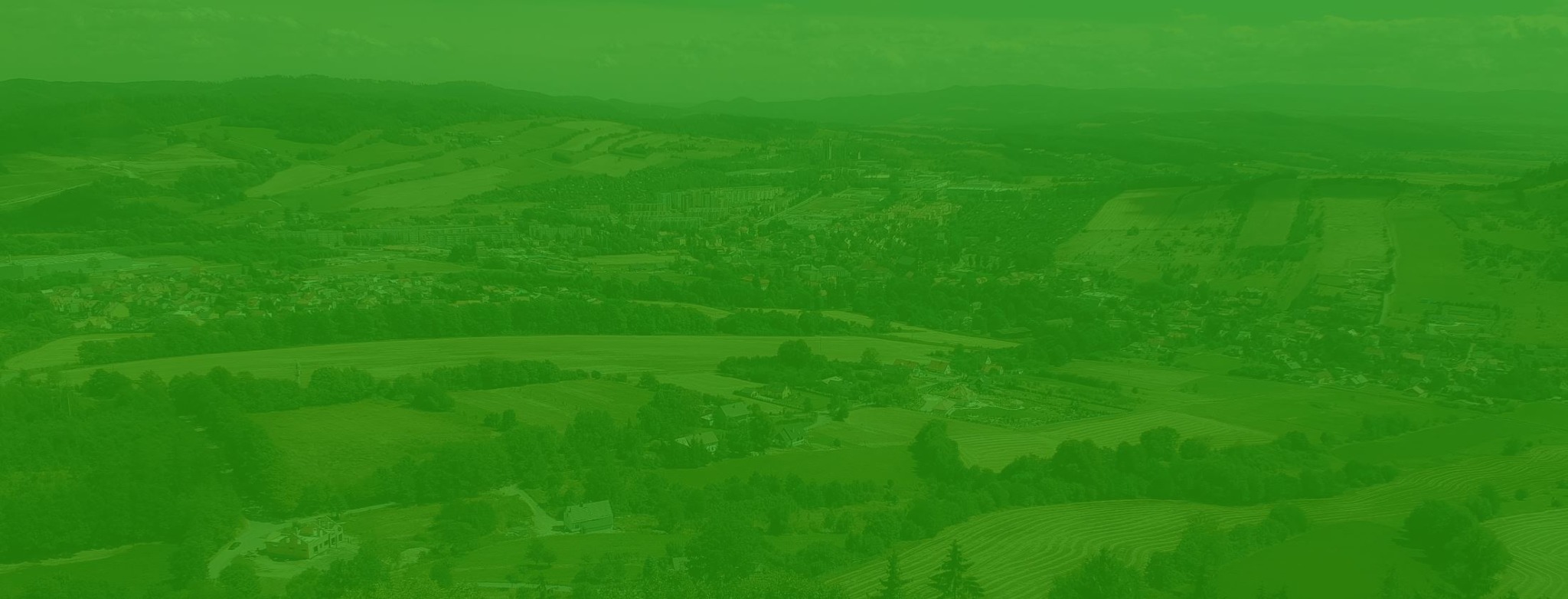 Panoramiczny widok na pagórkowaty krajobraz z widoczną zabudową mieszkalną i polami, wszystko utrzymane w odcieniach zieleni.