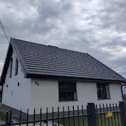 Dom jednorodzinny z ciemną dachówką karpiówką, świeżo otynkowaną elewacją w kolorze złamanej bieli i numerem '66' na rogu, widziany zza czarnego metalowego ogrodzenia na tle pochmurnego nieba.