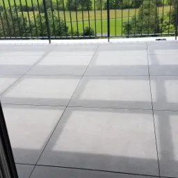 Szare, matowe płytki na balkonie z widokiem na zielony krajobraz za czarną metalową barierką, widoczne przez otwarte drzwi balkonowe.