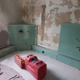 Wnętrze w trakcie remontu: ściany częściowo pokryte płytami gipsowo-kartonowymi z widocznymi instalacjami hydraulicznymi, surowa ściana z resztkami farby, na podłodze skrzynki narzędziowe Hilti...