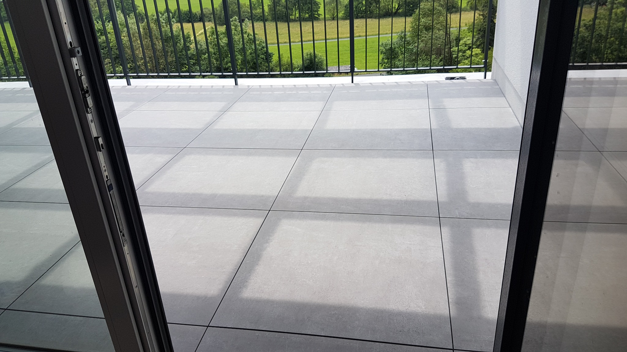 Szare, matowe płytki na balkonie z widokiem na zielony krajobraz za czarną metalową barierką, widoczne przez otwarte drzwi balkonowe.