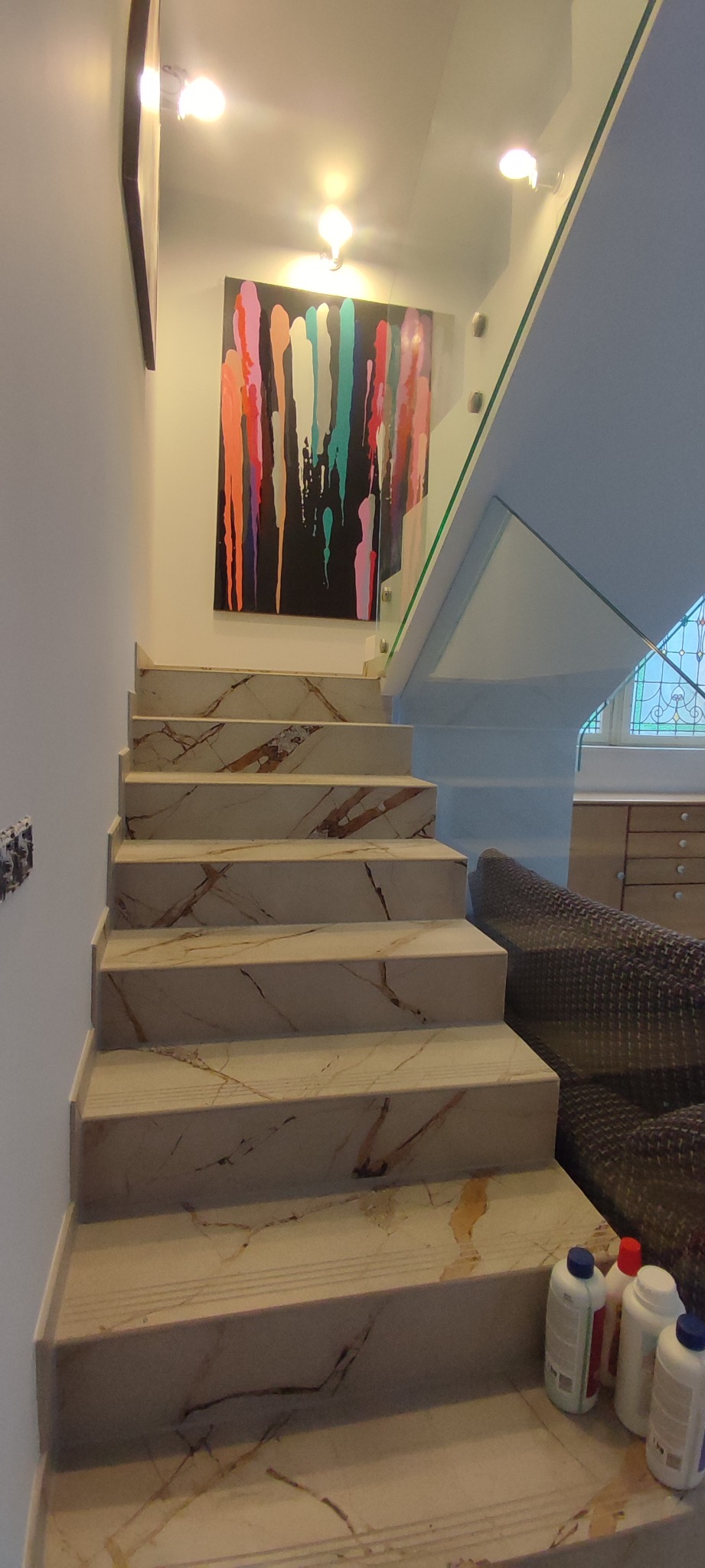 Marmurowe schody wewnętrzne z szklaną balustradą, oświetlone punktowymi lampami sufitowymi, z abstrakcyjnym obrazem wiszącym na ścianie i butelkami środków czyszczących na dolnym stopniu.
