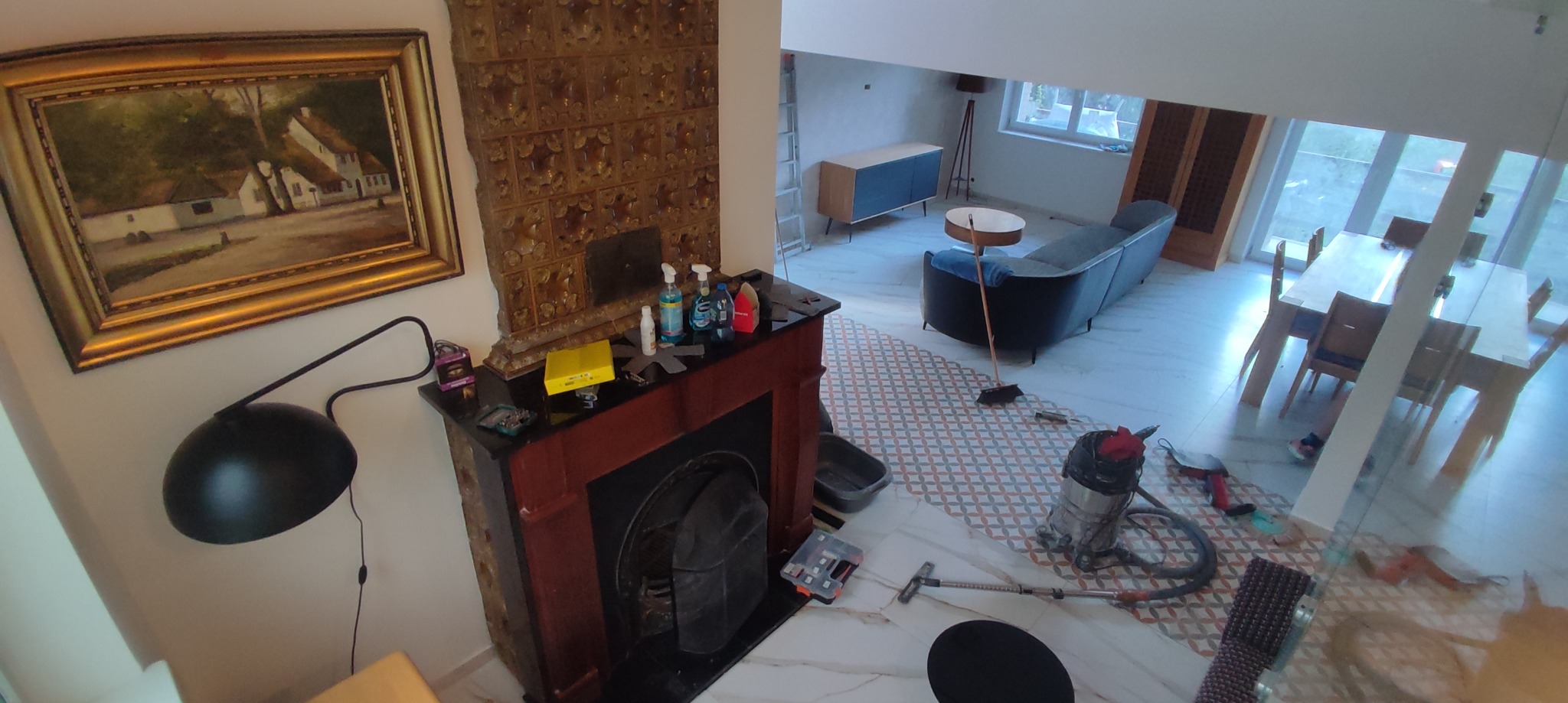 Wnętrze domu w trakcie prac wykończeniowych z widocznym kominkiem, nową podłogą z wzorzystymi płytkami, meblami i sprzętem budowlanym, uchwycone z góry.