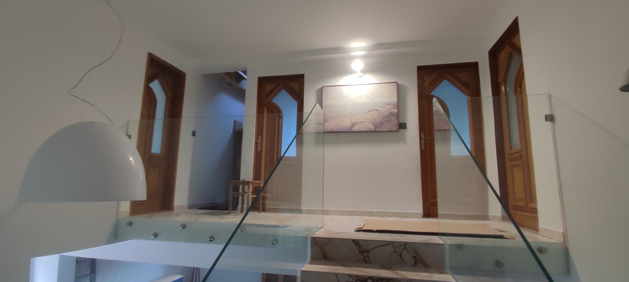 Nowoczesne wnętrze z przeszkloną balustradą, marmurowymi schodami i trzema drewnianymi drzwiami z geometrycznymi przeszkleniami. Na ścianie wisi obraz nad kartonem ochronnym.