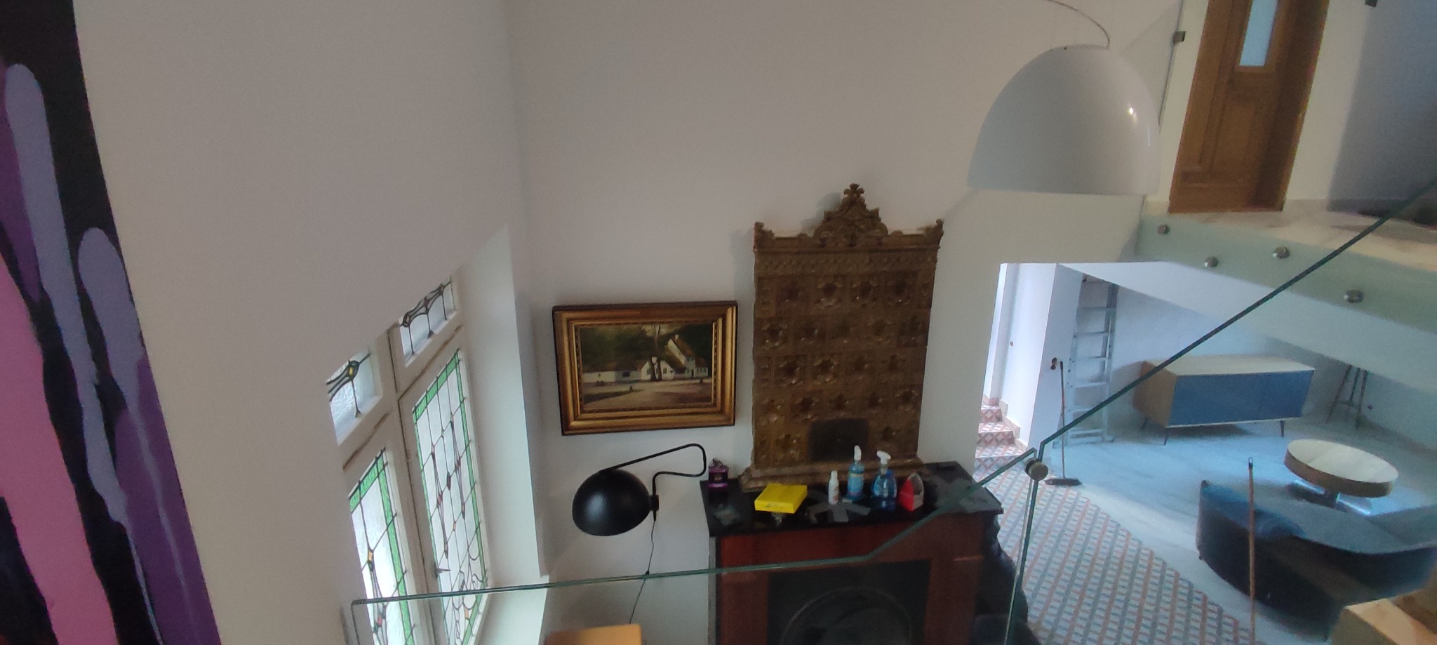 Wnętrze domu z widokiem na ozdobny piec kaflowy, obraz w złotej ramie, okno ze szprosami i nowoczesne schody ze szklaną balustradą.