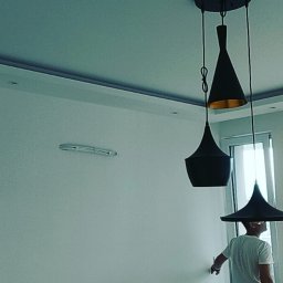 Ihor Voskoboinyk LedLamp V&B - Trzy czarne lampy wiszące o różnych kształtach, z elementami w kolorze złota w środku, na tle białej ściany w trakcie montażu przez mężczyznę w białej koszulce.