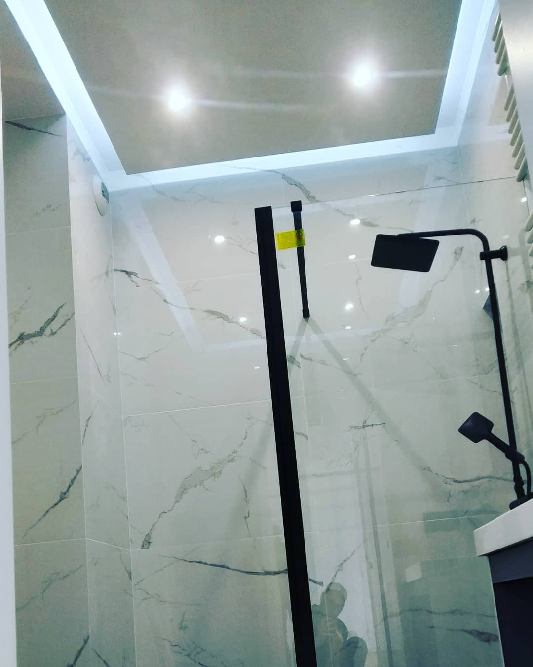 Nowoczesna kabina prysznicowa z czarnymi elementami armatury i szklaną ścianką, wykończona płytkami imitującymi marmur, oświetlona punktowymi lampami w podwieszanym suficie.