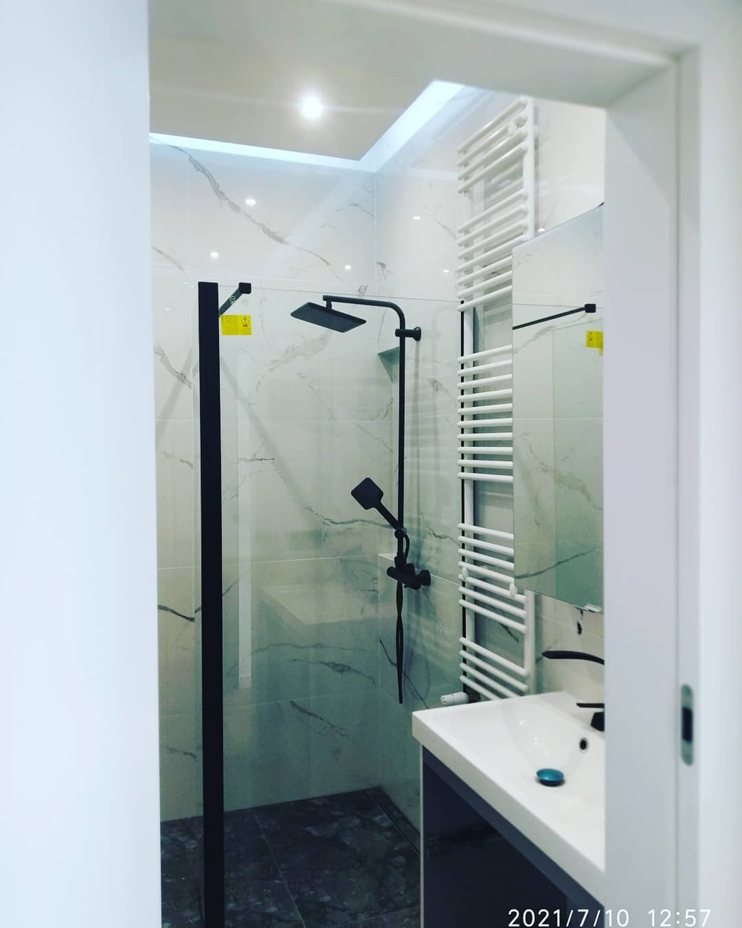 Nowoczesna łazienka z kabiną prysznicową typu walk-in z czarnymi elementami, marmurowe płytki na ścianach, grzejnik drabinkowy w kolorze białym i umywalka nablatowa.