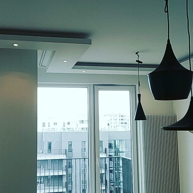 Nowoczesny salon w Warszawie z widokiem na miasto, minimalistyczne lampy wiszące i gładkie, jasne ściany z subtelnym oświetleniem sufitowym.