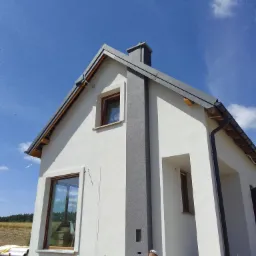 Dwukondygnacyjny dom z oknami w drewnianych ramach i szarą elewacją w trakcie prac wykończeniowych, widoczny pracownik w czerwonej koszulce przy elewacji, błękitne niebo z chmurami w tle.