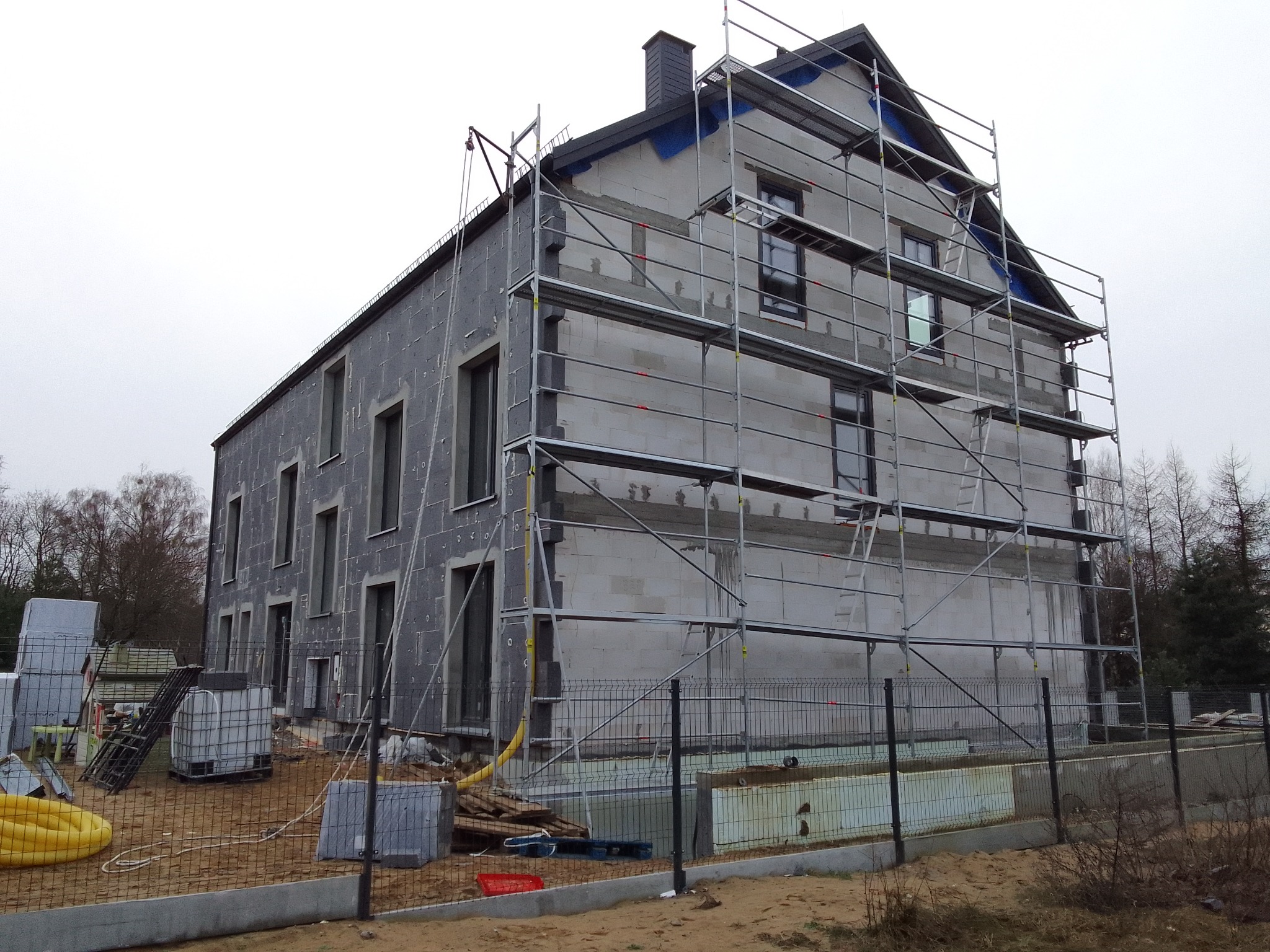 Dwukondygnacyjny dom w trakcie prac elewacyjnych, częściowo ocieplony szarym styropianem, z rusztowaniem na trzech ścianach, widoczne materiały budowlane na posesji.
