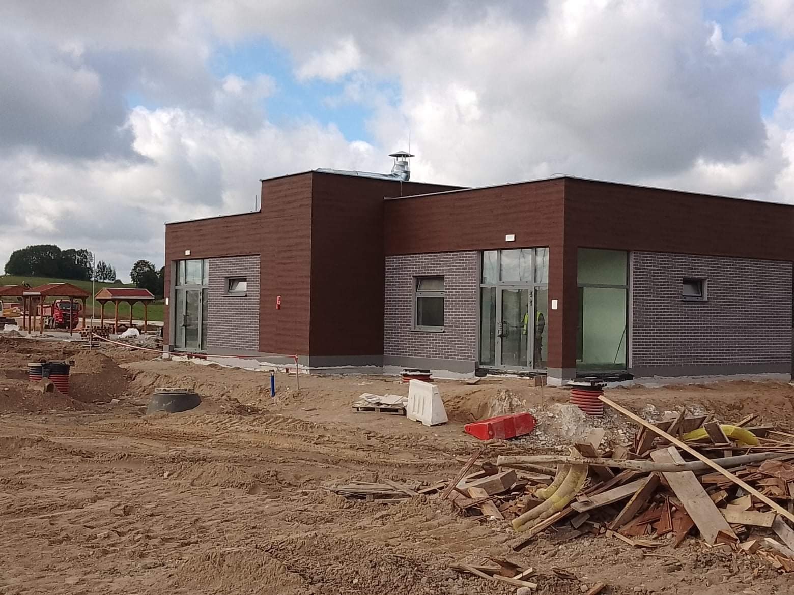 Nowoczesny budynek jednokondygnacyjny z elewacją z brązowego drewna i szarej cegły, widoczny w trakcie budowy, z rozkopanym terenem i stertą drewna na pierwszym planie.