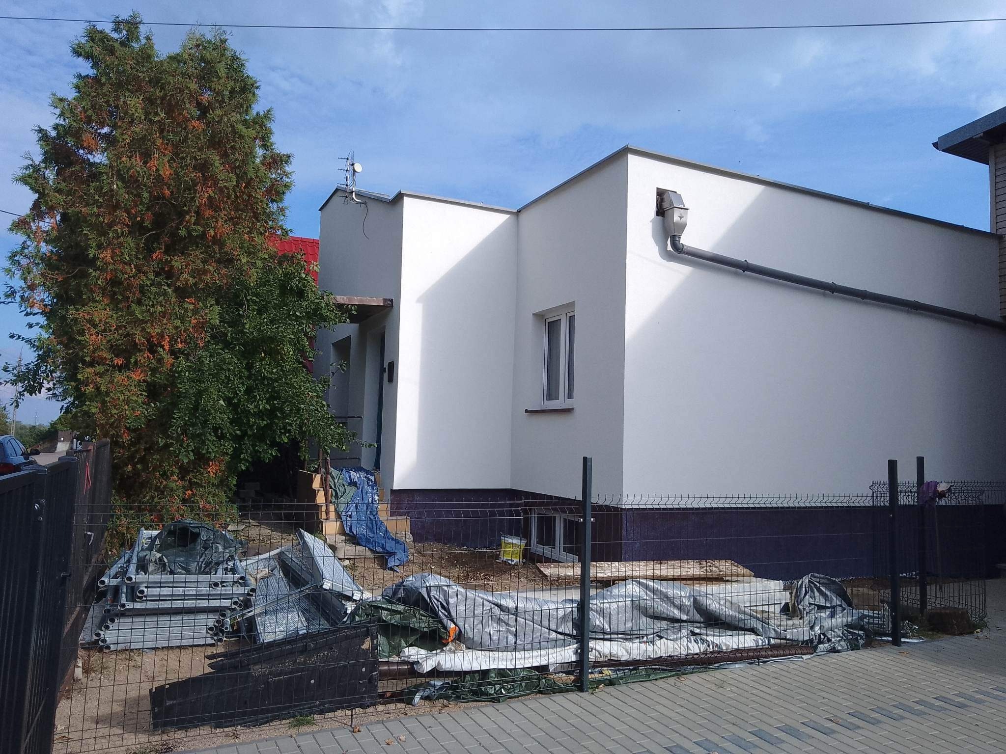 Świeżo otynkowany, biały budynek z widocznym systemem rynnowym i okienkiem, częściowo zasłonięty przez drzewo i ogrodzenie, za którym leżą zabezpieczone materiały budowlane i rusztowanie.