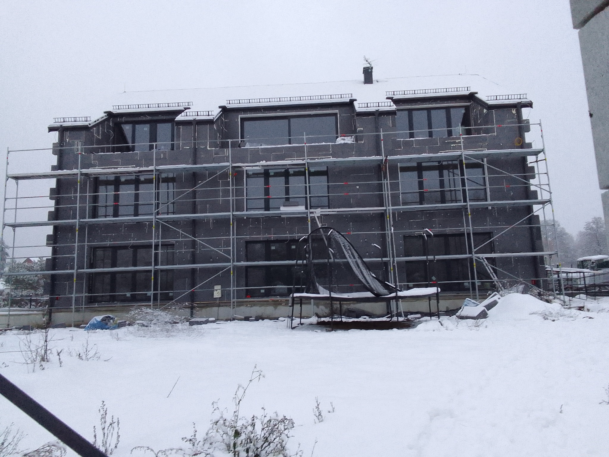 Wielopiętrowy budynek w trakcie ocieplania elewacji, pokryty szarym styropianem, otoczony rusztowaniem, zimowa sceneria z padającym śniegiem.