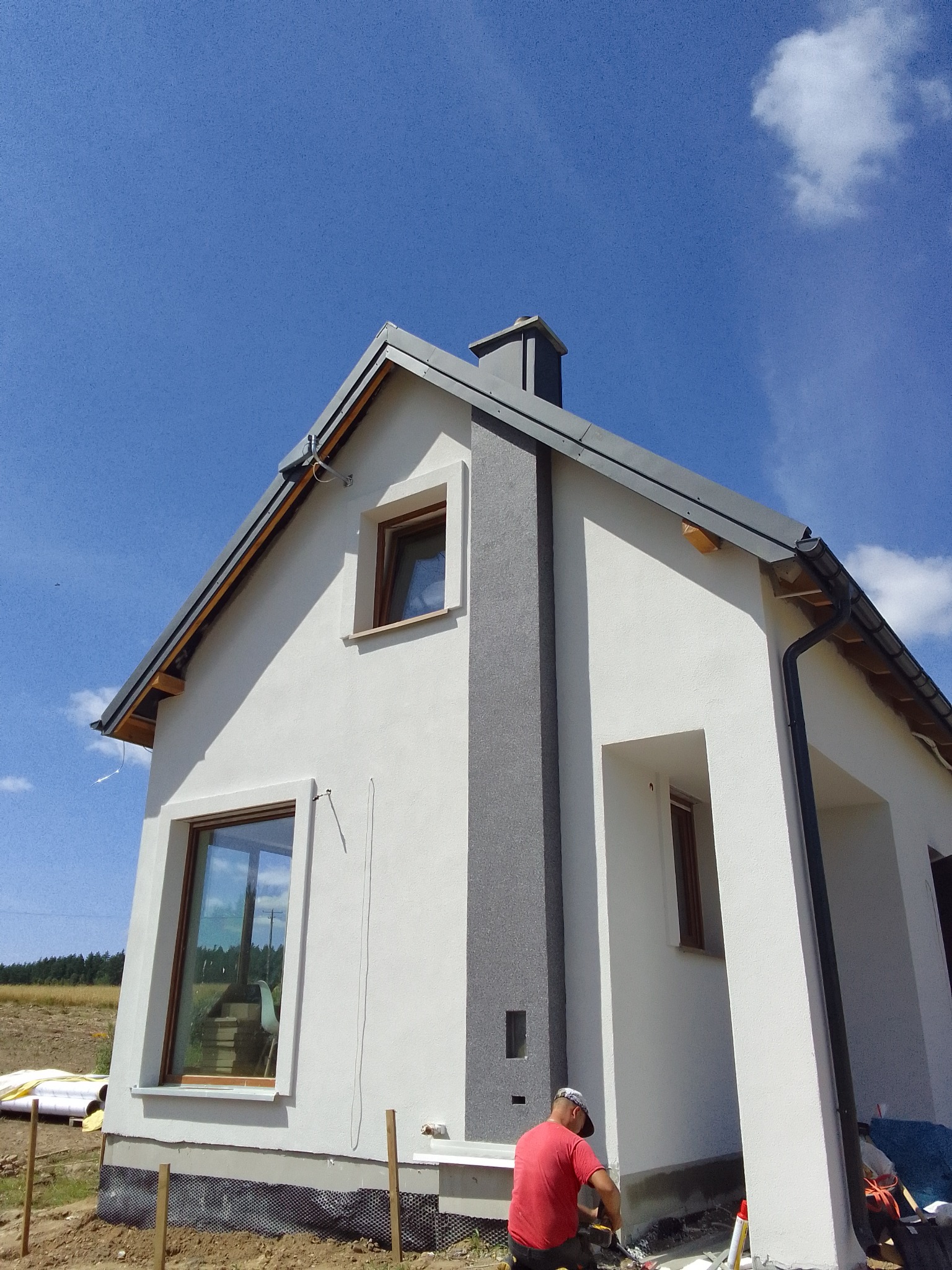 Dwukondygnacyjny dom z oknami w drewnianych ramach i szarą elewacją w trakcie prac wykończeniowych, widoczny pracownik w czerwonej koszulce przy elewacji, błękitne niebo z chmurami w tle.