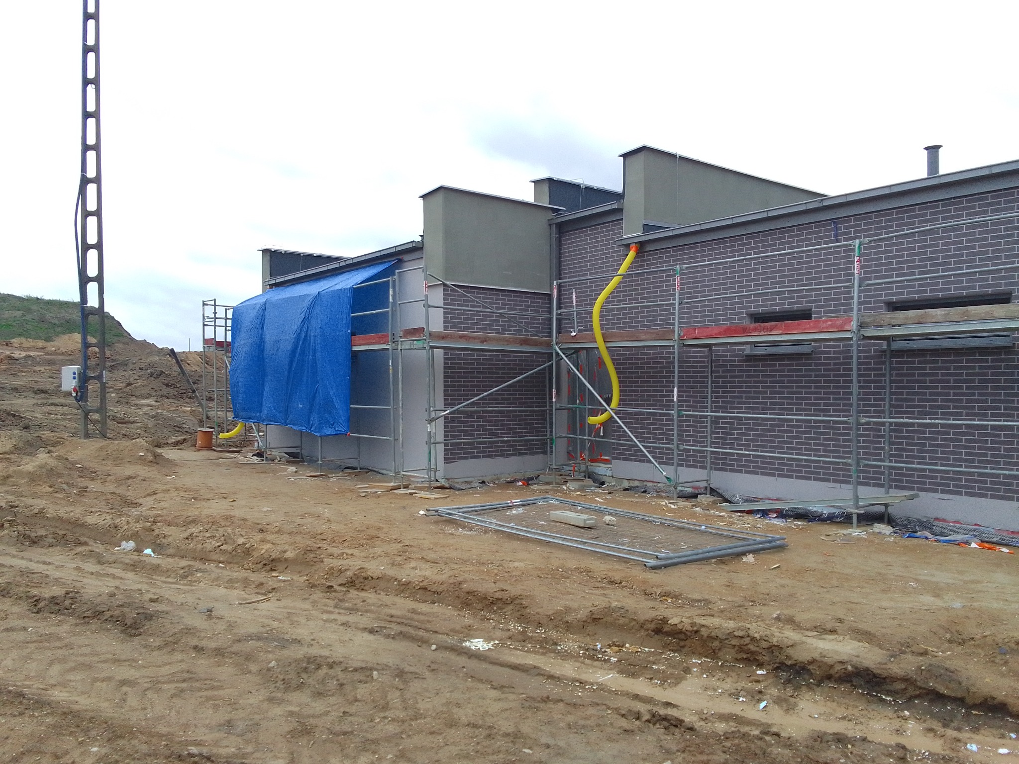 Budynek w trakcie prac elewacyjnych z rusztowaniem i żółtym przewodem wentylacyjnym, częściowo zasłonięty niebieską plandeką, teren budowy z ziemią i narzędziami.