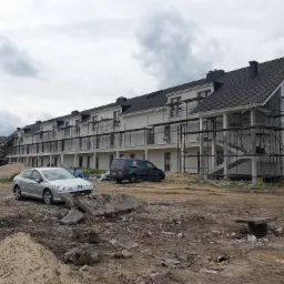 Wielorodzinny budynek w trakcie prac elewacyjnych, widoczne rusztowania, niezagospodarowany teren wokół, zaparkowane samochody.