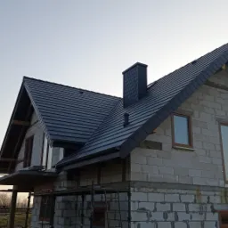 Dom w stanie surowym zamkniętym z czarną dachówką i kominem z tego samego materiału, widoczne rusztowania i okna z brązowymi ramami, w tle drzewa i turbiny wiatrowe.