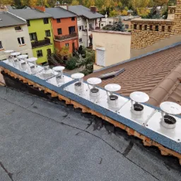 Szereg nowych, srebrnych zakończeń kominowych z daszkami, zamontowanych na krawędzi dachu pokrytego papą, z widocznymi ceglanymi elementami konstrukcyjnymi i fragmentem brązowej dachówki w tle,...