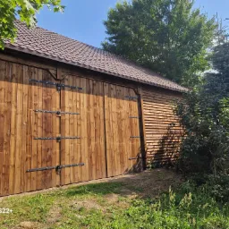 Nowy, brązowy dach z blachodachówki trapezowej nad drewnianym budynkiem gospodarczym z dużymi, dwuskrzydłowymi wrotami, widok w słoneczny dzień.