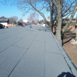 Ekipa dekarzy podczas układania papy termozgrzewalnej na płaskim dachu, widoczny cylinder z gazem i narzędzia, w tle zabudowania gospodarcze i drzewa bez liści.