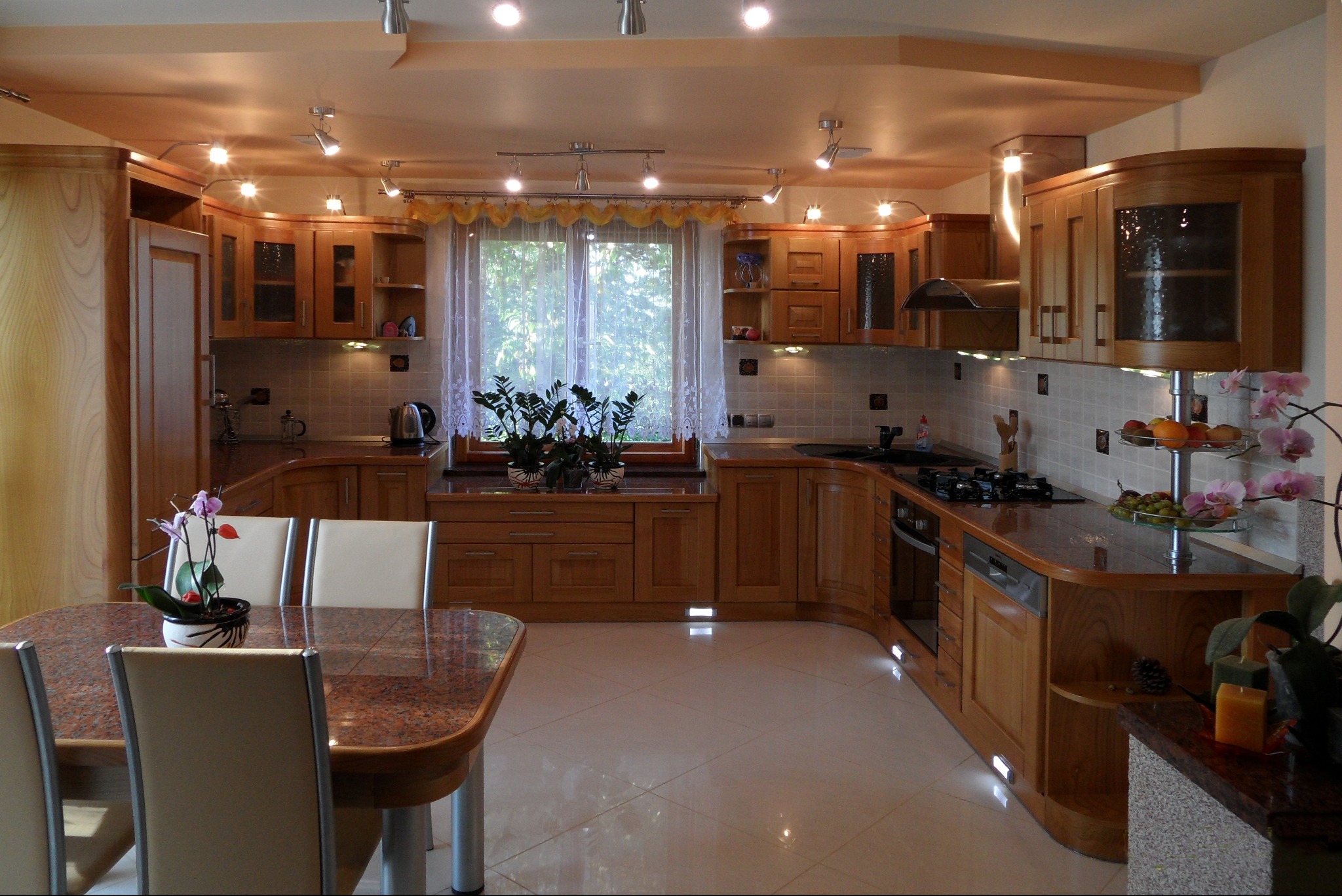 Jasna, drewniana kuchnia z blatem z granitu, oświetleniem punktowym i roślinami w oknie, widok z perspektywy stołu jadalnego z kwiatem.