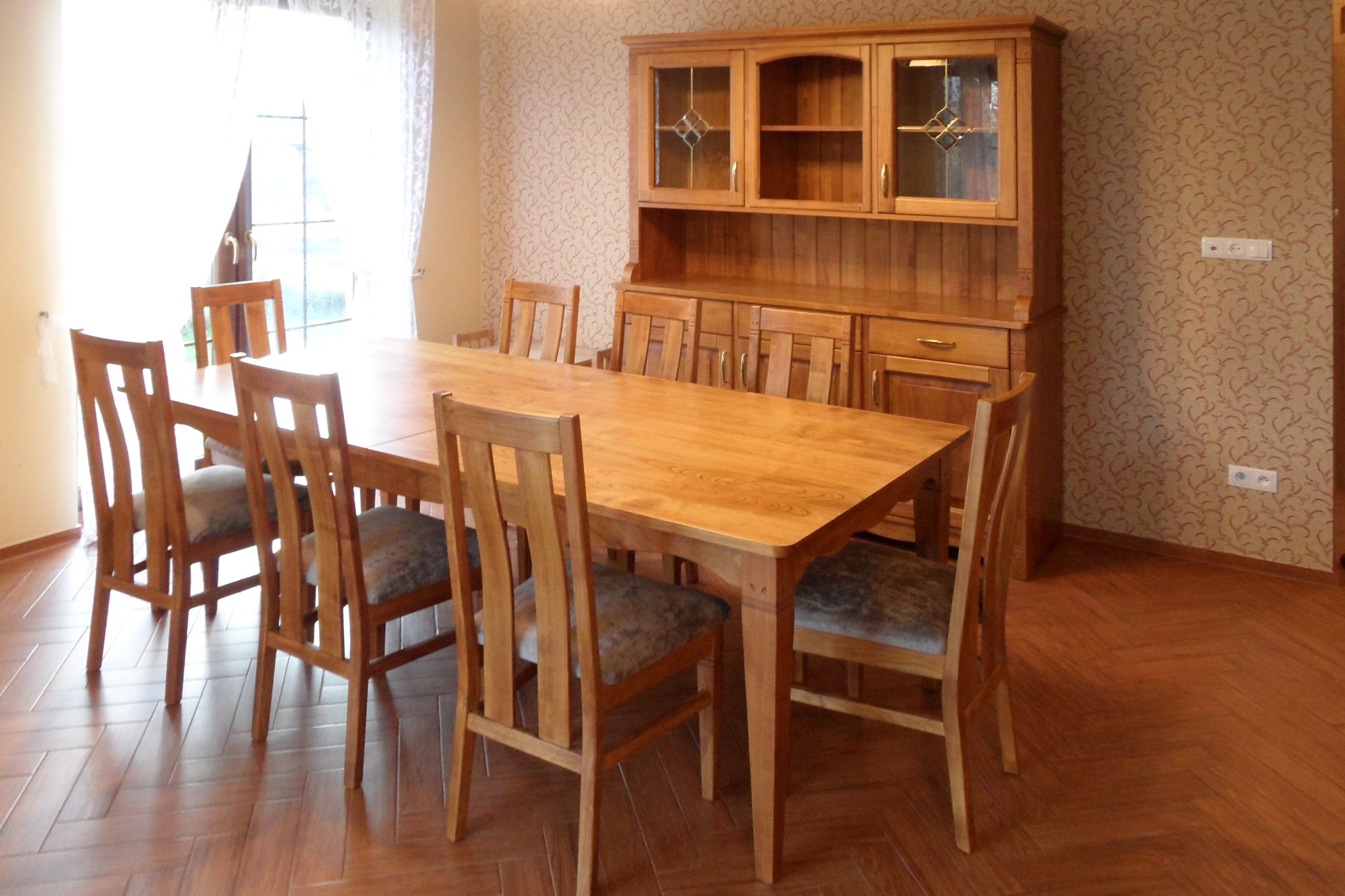 Drewniany stół jadalny z sześcioma krzesłami i kredensem w tle, na podłodze parkiet ułożony w jodełkę, wzorzysta tapeta na ścianie.