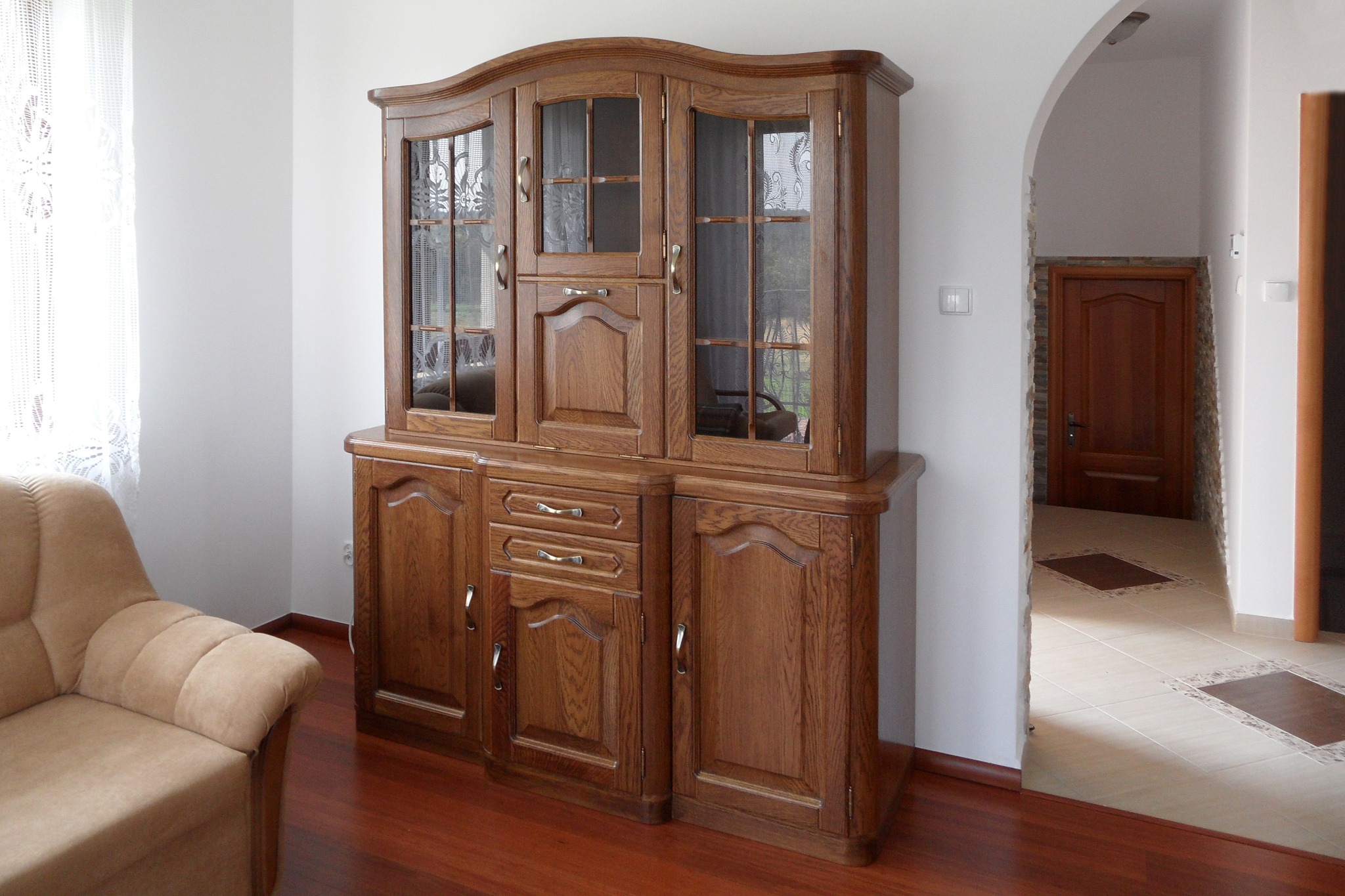 Rustykalny kredens z drewna dębowego, z przeszkloną nadstawką i szafkami, widoczny w jasnym wnętrzu z drewnianą podłogą i beżowym fotelem