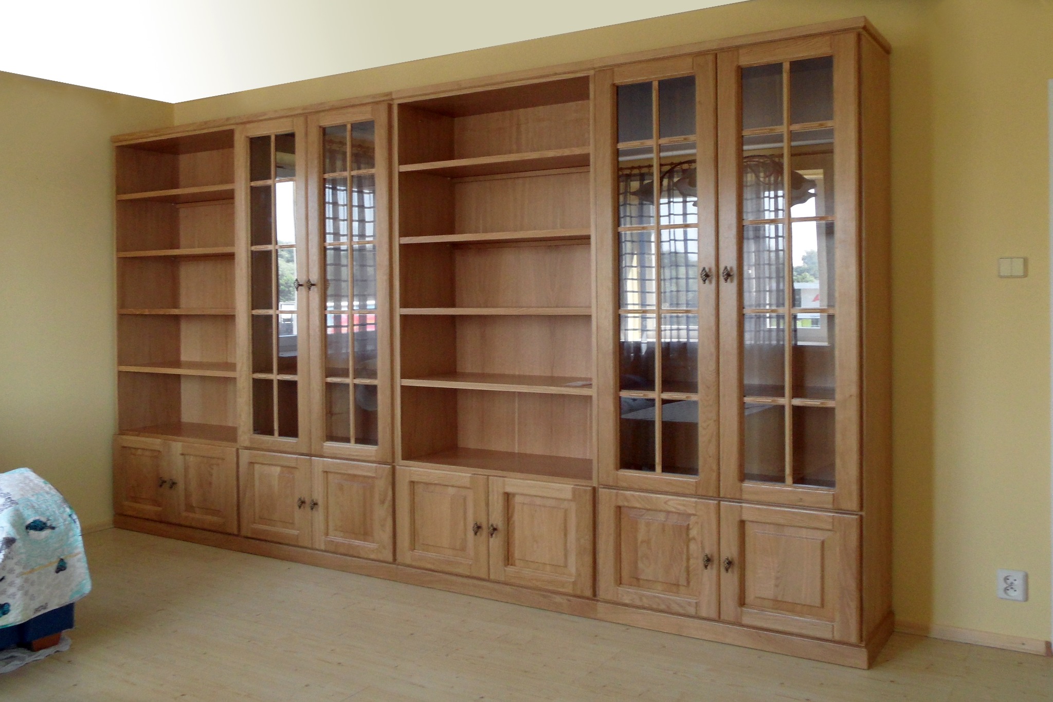 Drewniana zabudowa meblowa z przeszklonymi witrynami i szafkami dolnymi, w jasnym odcieniu drewna, dopasowana do wnęki pomieszczenia z parkietem.