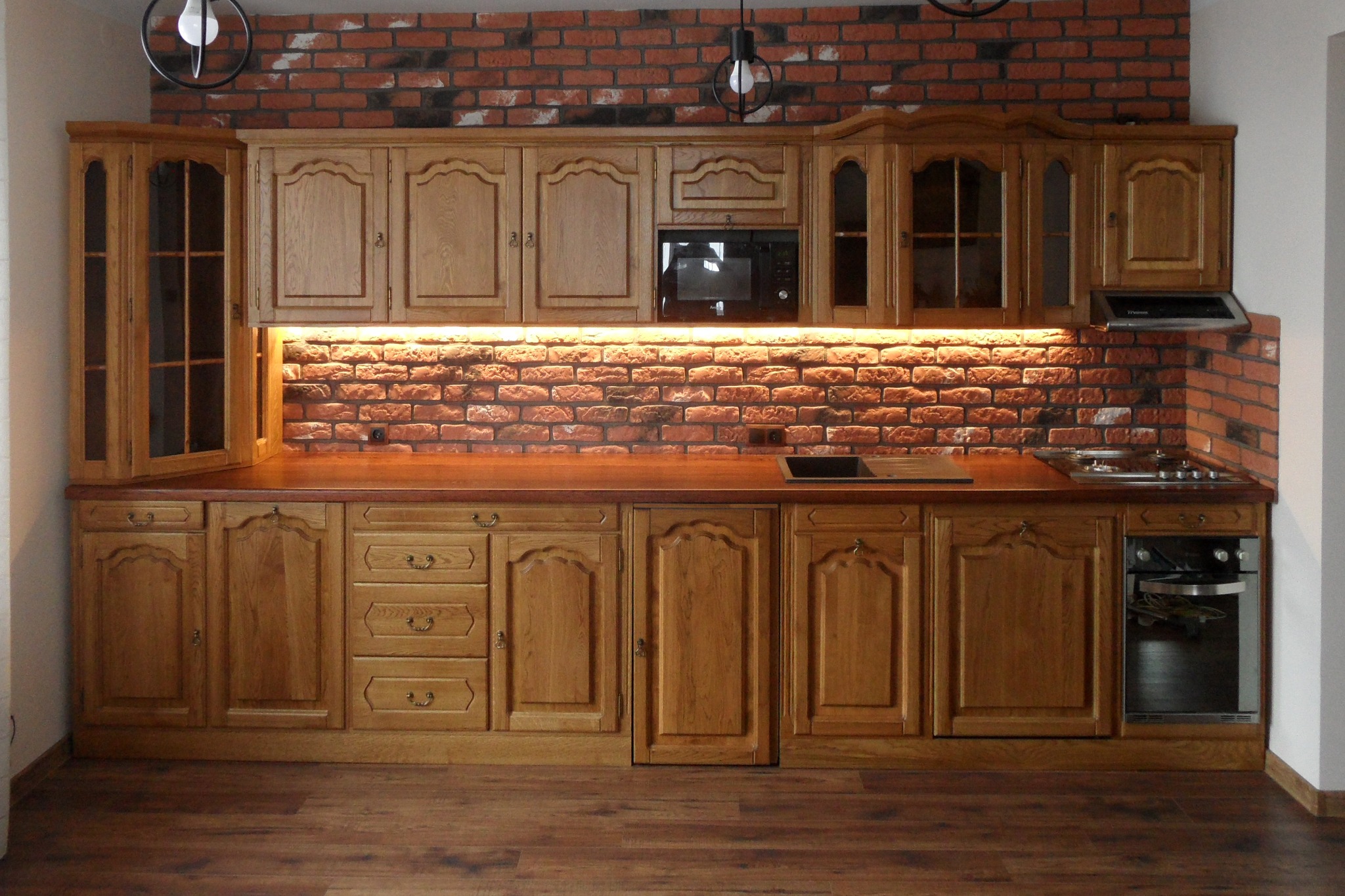 Rustykalna kuchnia z drewnianymi szafkami, blatem w ciepłym odcieniu i ścianą z czerwonej cegły podświetloną ledami, widoczne oświetlenie sufitowe w stylu industrialnym.