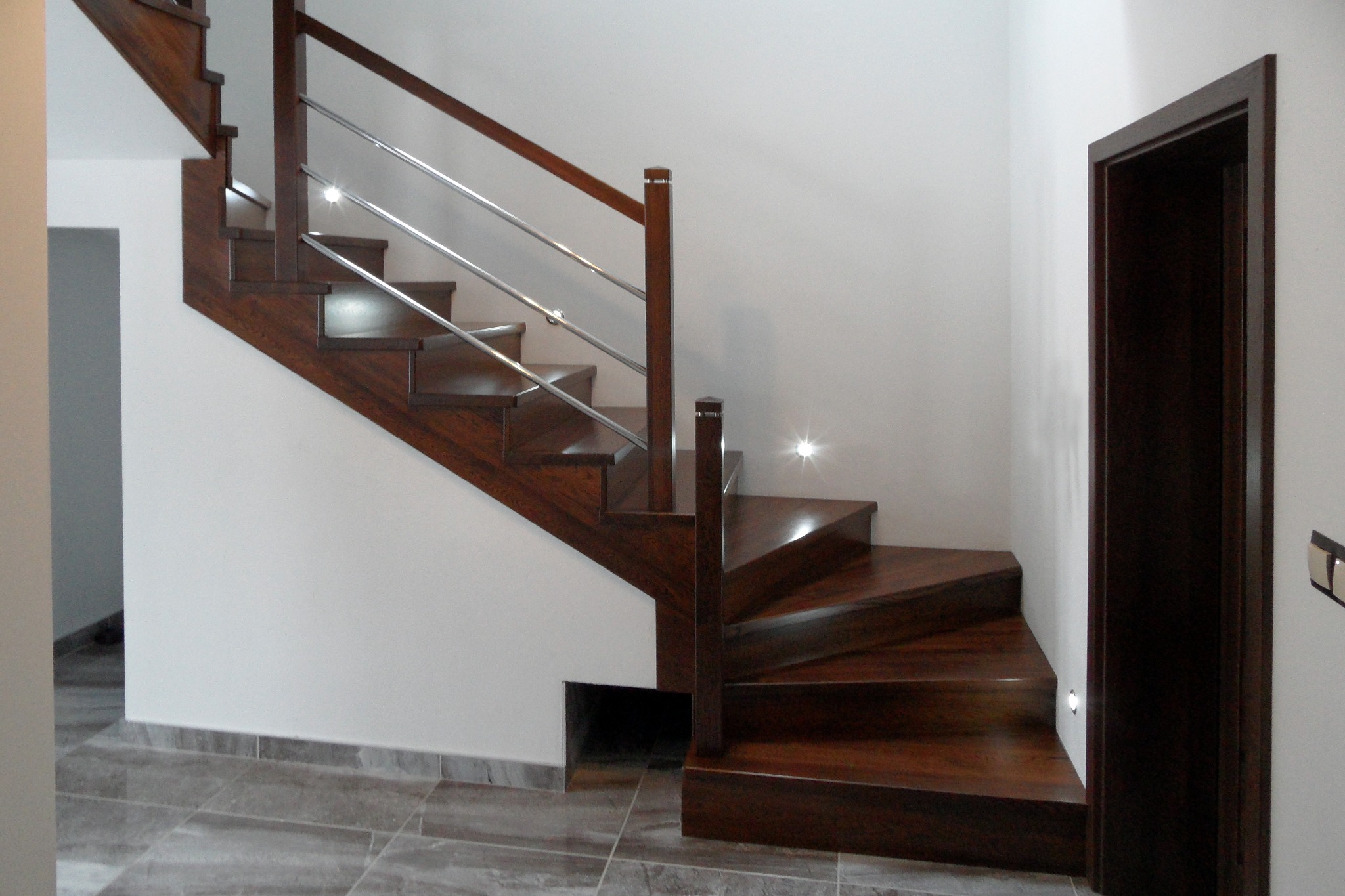 Nowoczesne, drewniane schody z metalową balustradą, oświetleniem LED na stopniach i ciemnym wykończeniem, kontrastujące z białymi ścianami i szarą podłogą.